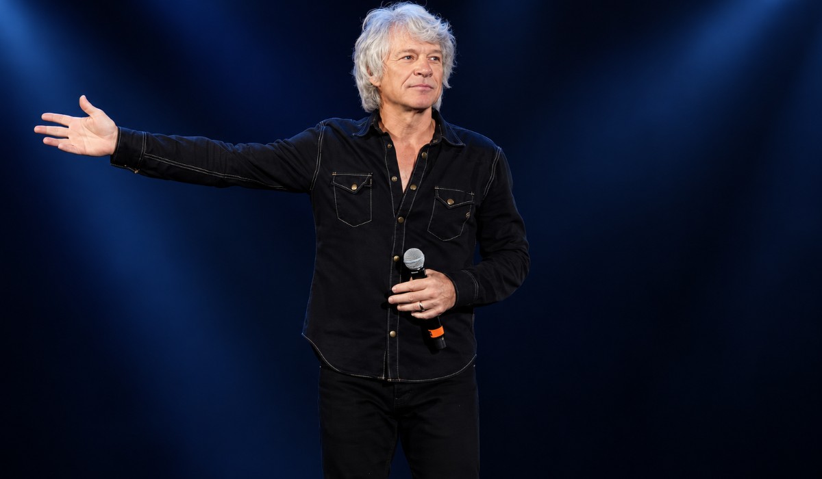 Jon Bon Jovi with silver hair and a black shirt holds a microphone and extends his arm against a dark blue concert background.