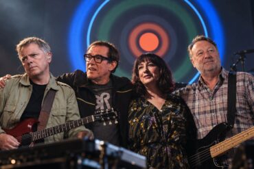 Joy Division - New Order members standing together on stage with the Blue Monday artwork in the background.