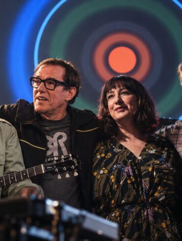 Joy Division and New Order Enter the Rock Hall Together - Here Is The Story Behind Their Shared History Joy Division - New Order members standing together on stage with the Blue Monday artwork in the background.