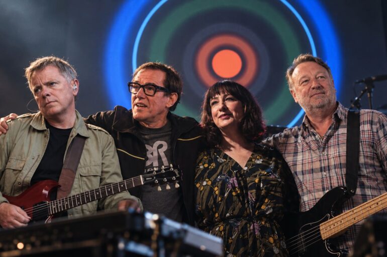 Joy Division - New Order members standing together on stage with the Blue Monday artwork in the background.