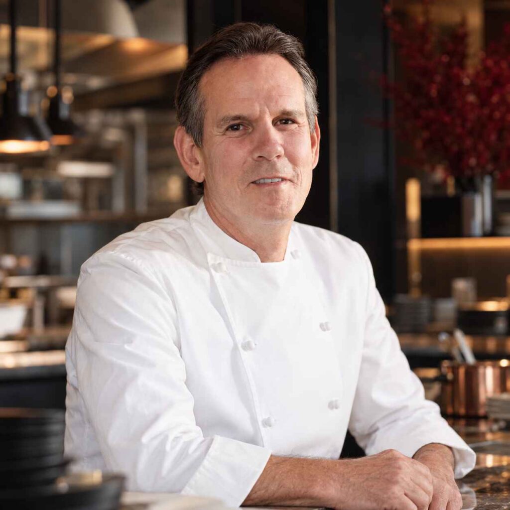 7 Famous Chefs Who Started Their Careers Washing Dishes Chef Thomas Keller wearing a white chef's jacket in a warmly lit restaurant kitchen.
