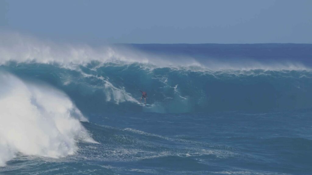 Surfer riding a towering ocean wave with powerful whitewater breaking behind.