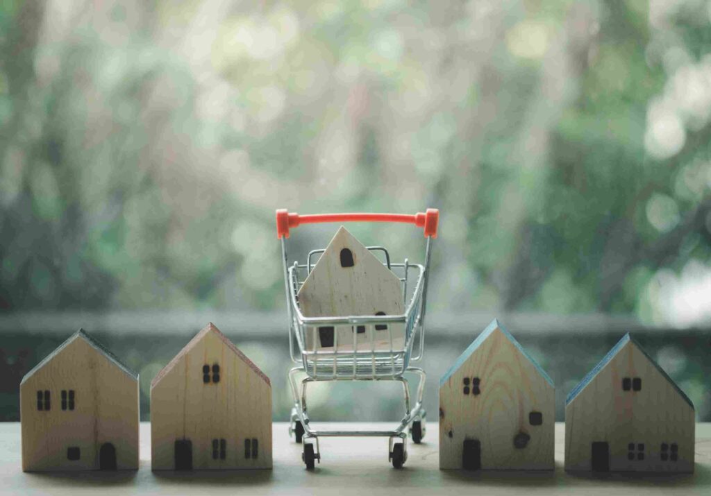 Seattle Housing Market 2026, Is Now a Better Time to Buy a House? Miniature wooden houses surrounding a shopping cart, representing homebuyers comparing properties.